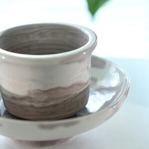 May include: A handmade ceramic cup and saucer set. The cup is cylindrical with a slightly flared rim, featuring bands of white, gray, and brown. The saucer is a matching round shape. The set is glazed with a glossy finish.