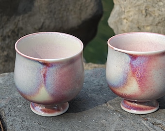 May include: Two handmade ceramic cups with a footed base. The cups have a white interior and a pink and purple exterior glaze. The cups are sitting on a gray stone surface.