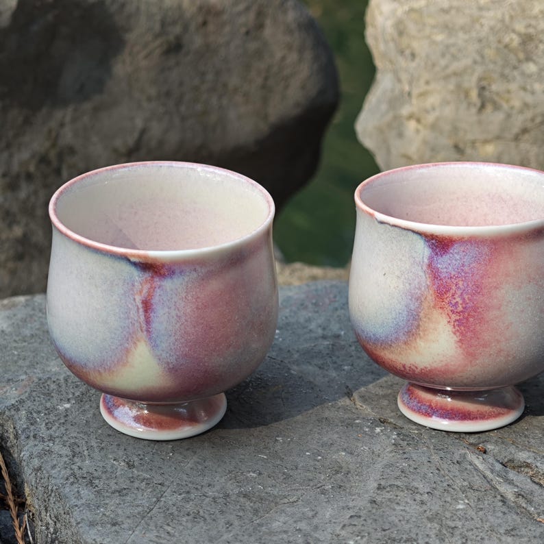 May include: Two handmade ceramic cups with a footed base. The cups have a white interior and a pink and purple exterior glaze. The cups are sitting on a gray stone surface.