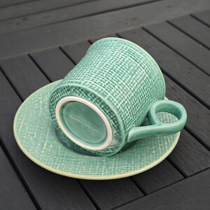 May include: A teal ceramic teacup and saucer set. The cup has a rounded handle and a textured woven pattern. The saucer is round and matches the cup's color and texture. The bottom of the cup has the text "Made with Love, Handle with Care."