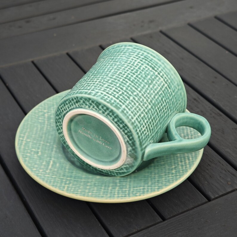 May include: A teal ceramic teacup and saucer set. The cup has a rounded handle and a textured woven pattern. The saucer is round and matches the cup's color and texture. The bottom of the cup has the text "Made with Love, Handle with Care."