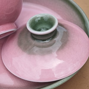 Puede incluir: Una tapa de cer&aacute;mica con un acabado de esmalte rosa brillante con efecto craquelado y un pomo central que presenta un interior verde salvia y una zona de transici&oacute;n gris oscuro. La tapa es circular con un borde liso y redondeado.