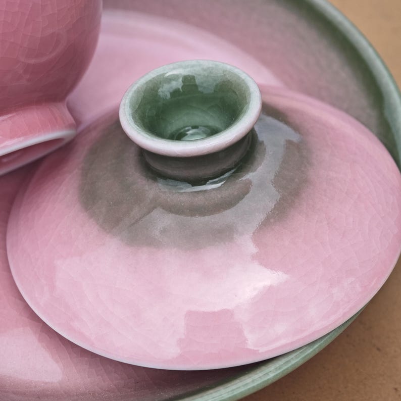 Puede incluir: Una tapa de cer&aacute;mica con un acabado de esmalte rosa brillante con efecto craquelado y un pomo central que presenta un interior verde salvia y una zona de transici&oacute;n gris oscuro. La tapa es circular con un borde liso y redondeado.