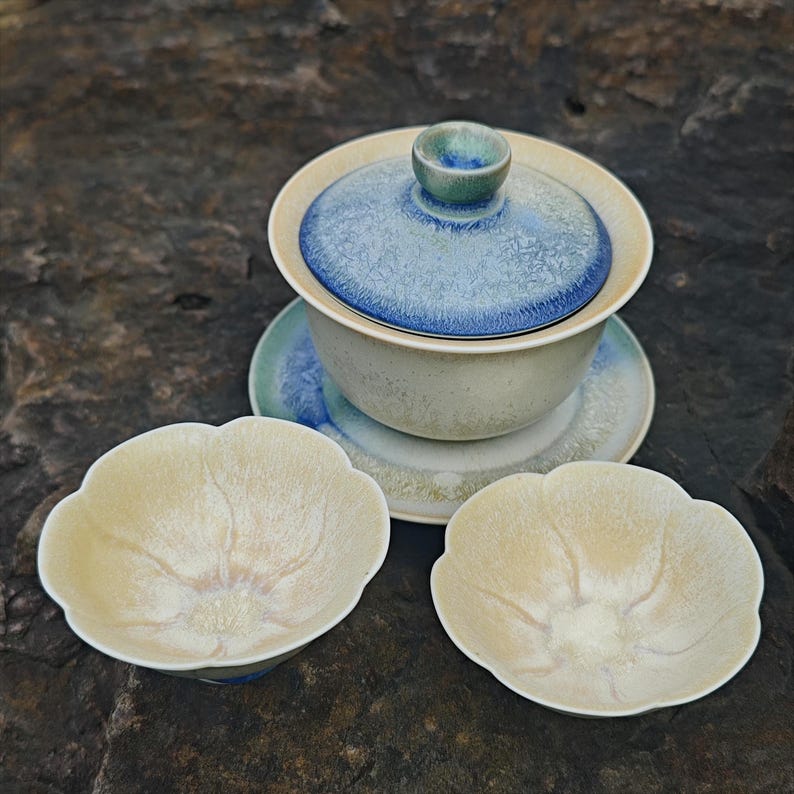 May include: A ceramic tea set with a light blue and cream color scheme. The set includes a lidded gaiwan, a saucer, and two small, petal-shaped teacups. The gaiwan has a blue-patterned lid and a matching handle. The teacups have a cream-colored interior.