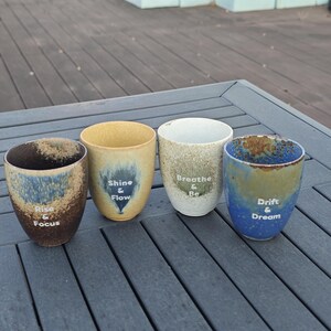 Puede incluir: Cuatro tazas de cer&aacute;mica con dise&ntilde;os &uacute;nicos y frases inspiradoras. Las tazas presentan una variedad de colores, incluyendo marr&oacute;n, azul, amarillo y blanco. Cada taza tiene una frase diferente: "Rise & Focus", "Shine & Flow", "Breathe & Be" y "Drift & Dream".
