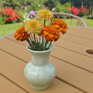 Puede incluir: Un jarr&oacute;n verde claro con un dise&ntilde;o floral contiene un ramo de flores naranjas y amarillas. Las flores est&aacute;n en plena floraci&oacute;n, con colores vibrantes. El jarr&oacute;n est&aacute; sobre una mesa de madera marr&oacute;n, con un fondo borroso de follaje verde.