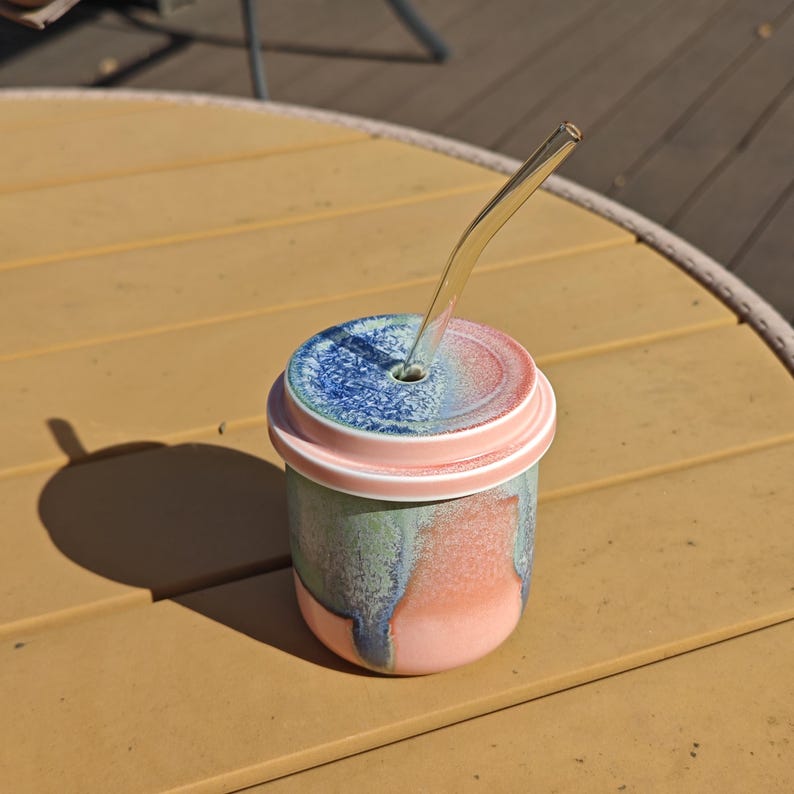 Pu&ograve; includere: Un bicchiere in ceramica con coperchio e cannuccia di vetro. Il bicchiere ha una smaltatura rosa, verde e blu. Il coperchio &egrave; rosa e blu e la cannuccia &egrave; trasparente. Il bicchiere &egrave; su un tavolo giallo. Altezza circa 10 cm.