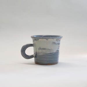 May include: A handcrafted ceramic mug with a curved handle. The mug displays a gradient of blue and white tones, with a darker blue base and rim. The mug is designed for hot drinks.