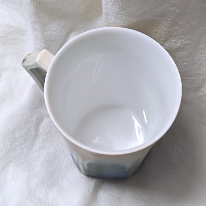 May include: A white ceramic mug with a geometric design. The mug has a white interior and a handle with a light beige and gray color. The exterior of the mug transitions from white to a light blue color.