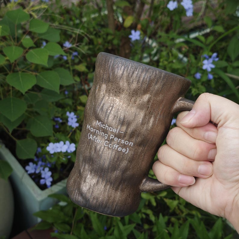 Puede incluir: Una taza cil&iacute;ndrica de cer&aacute;mica con un acabado de bronce met&aacute;lico texturizado que se asemeja a la corteza de un &aacute;rbol. La taza presenta un texto impreso en blanco: Michael - Morning Person (After Coffee). Tiene un asa resistente en el lado.