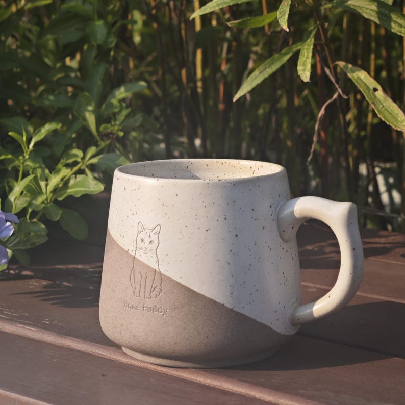 May include: A two-toned ceramic mug with a speckled cream upper section and a solid light brown lower section. The mug features a line drawing of a cat and the text "home buddy" embossed on the front, along with a curved handle on the right side.