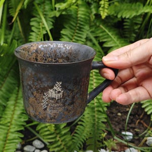 May include: A dark ceramic mug with a handle, showing a faded white graphic. The mug has a textured, speckled appearance with brown and black tones. The background is a lush green fern. The mug is likely around 10 cm tall.