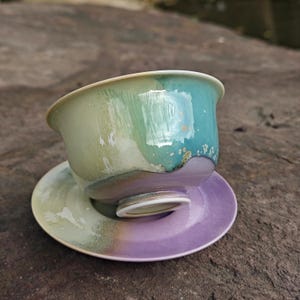 May include: A ceramic teacup and saucer set. The teacup is a blend of green and blue hues, with a white rim. The saucer is a gradient of purple and green. The set is designed for tea or coffee.
