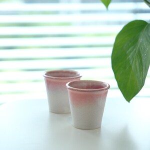 May include: Two ceramic cups with a gradient design, transitioning from a soft pink at the rim to a creamy white at the base. The cups are cylindrical with a slightly flared rim, set on a white surface. The background includes a window with blinds and a green plant.