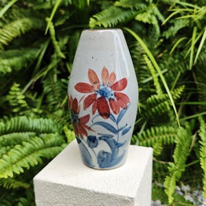 May include: A ceramic vase with a light gray background and a floral design. The flowers are red with blue centers and blue stems and leaves. The vase is approximately 6 inches tall.