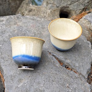 May include: Two ceramic tea cups with a cream-colored interior and a blue band around the base. The cups have a gold rim and sit on a gray rock surface. The cups are designed for tea or other beverages.