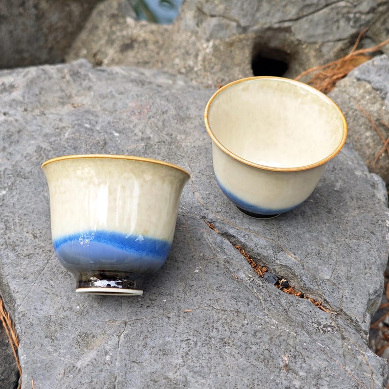 May include: Two ceramic tea cups with a cream-colored interior and a blue band around the base. The cups have a gold rim and sit on a gray rock surface. The cups are designed for tea or other beverages.