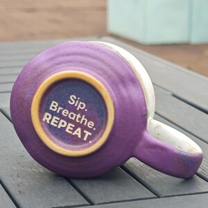 Op de afbeelding: Een paarse keramische mok met een witte rand en oor, van onderaf gezien. De bodem van de mok heeft de tekst "Sip. Breathe. REPEAT." in het wit, binnen een goudkleurige cirkel.