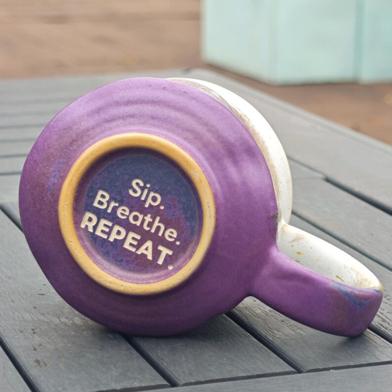 Op de afbeelding: Een paarse keramische mok met een witte rand en oor, van onderaf gezien. De bodem van de mok heeft de tekst "Sip. Breathe. REPEAT." in het wit, binnen een goudkleurige cirkel.