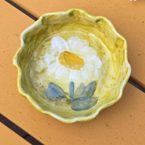 May include: A small, yellow ceramic bowl with a scalloped edge. The bowl features a hand-painted daisy design in the center, with white petals and a yellow center. Blue leaves and stem are also painted on the bowl.