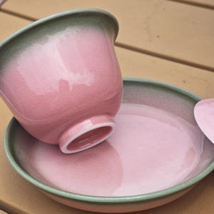 Puede incluir: Un cuenco de t&eacute; de cer&aacute;mica y su plato a juego con un esmalte rosa brillante y un fino patr&oacute;n de grietas. Los bordes de ambas piezas tienen un degradado en verde salvia apagado. El cuenco est&aacute; inclinado, apoyado parcialmente en el plato.