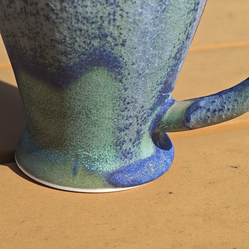 May include: A handmade ceramic mug with a speckled glaze in shades of blue and green. The mug features a curved handle and a wide base with a white rim. The mug's surface has a textured appearance.