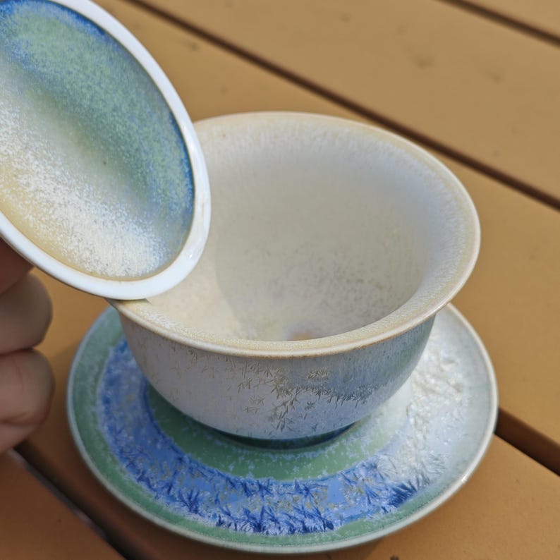 May include: A ceramic tea set with a cup, saucer, and lid. The cup and saucer have a blue, green, and white color scheme. The lid is white with blue and green accents. The set is on a wooden surface.