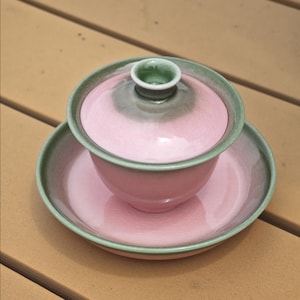 May include: A ceramic gaiwan tea set consisting of a bowl, a lid, and a saucer. The pieces feature a soft pink glaze in the center that transitions into a muted sage green gradient along the edges and rims. The lid has a small, raised knob.