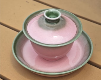 May include: A ceramic gaiwan tea set consisting of a bowl, a lid, and a saucer. The pieces feature a soft pink glaze in the center that transitions into a muted sage green gradient along the edges and rims. The lid has a small, raised knob.