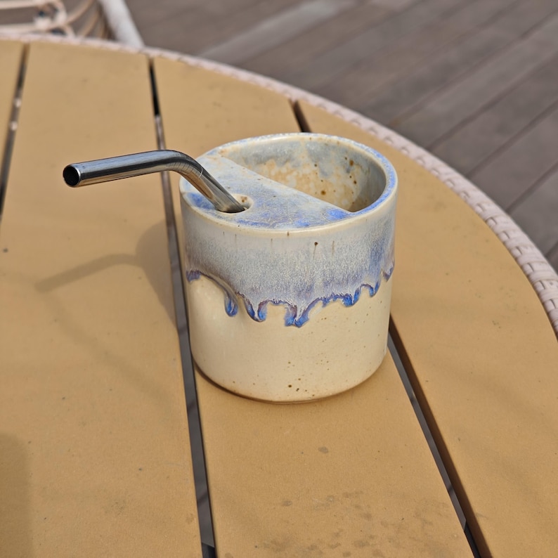May include: A cylindrical ceramic cup with a cream-colored base and a blue drip glaze pattern near the rim. The cup features a built-in horizontal shelf with a circular opening, holding a bent stainless steel metal straw for drinking beverages.