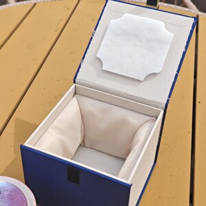 K&ouml;nnte beinhalten: Eine quadratische, offene Geschenkbox mit tiefblauer Au&szlig;enseite und cremefarbenem Stofffutter. Die Innenseite des Deckels ist mit einer wei&szlig;en Papiereinlage mit gewelltem Rand versehen. Die Box hat einen Klappdeckel und einen schwarzen Verschluss.