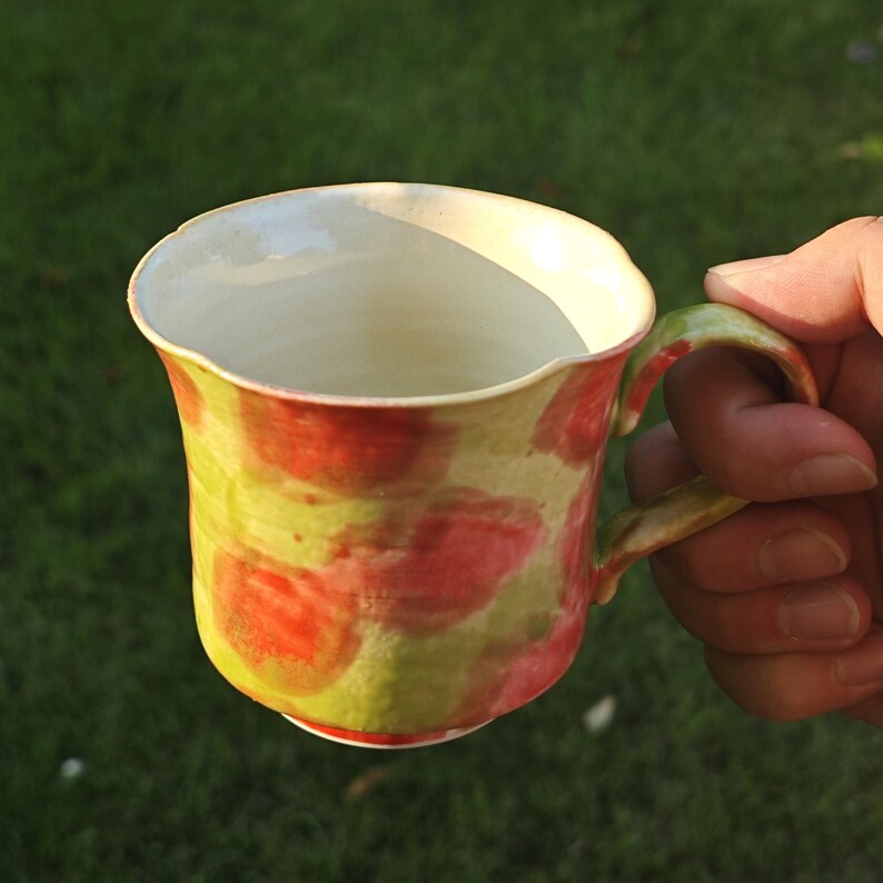 May include: A handmade ceramic mug with a unique design. The mug features a vibrant color palette of red and green blotches over a cream-colored base. The handle and rim are also painted in complementary colors. The mug is held against a green background.