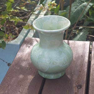 Puede incluir: Un jarr&oacute;n de cer&aacute;mica verde p&aacute;lido con cuerpo redondo y borde acampanado. El jarr&oacute;n tiene un patr&oacute;n texturizado y cristalino y est&aacute; sobre una superficie de madera. El jarr&oacute;n mide aproximadamente 18 cm de alto.