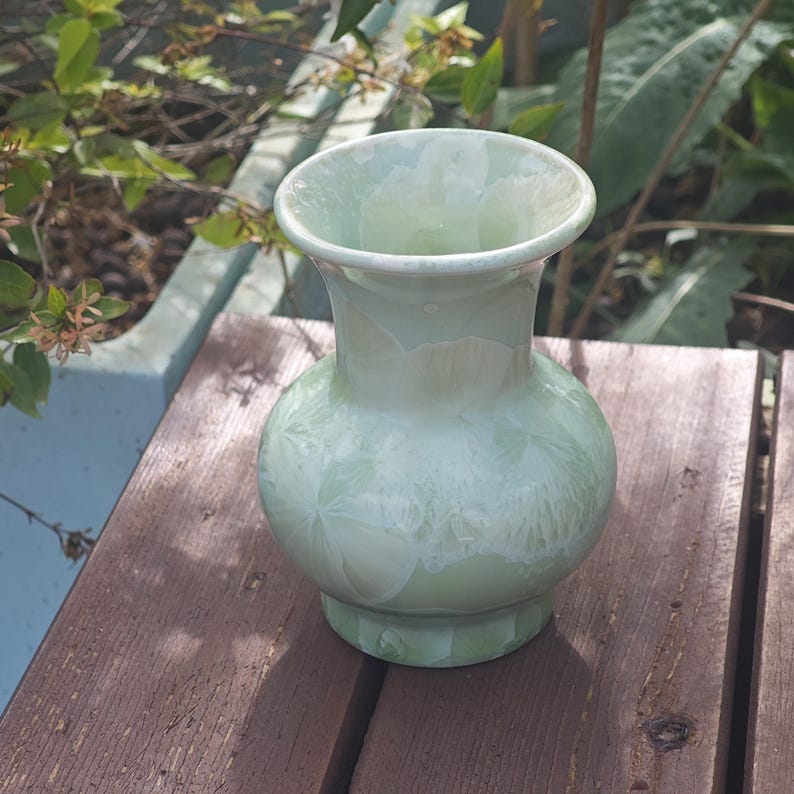 Puede incluir: Un jarr&oacute;n de cer&aacute;mica verde p&aacute;lido con cuerpo redondo y borde acampanado. El jarr&oacute;n tiene un patr&oacute;n texturizado y cristalino y est&aacute; sobre una superficie de madera. El jarr&oacute;n mide aproximadamente 18 cm de alto.