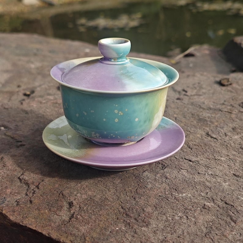 May include: A ceramic tea set with a lid, cup, and saucer. The set features a gradient of colors, including blue, purple, and green. The cup and saucer are round, and the lid has a small handle. The set is designed for tea.