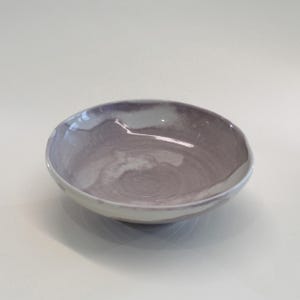 May include: A ceramic bowl with a light purple and white swirl pattern. The bowl has a rounded shape with a slightly uneven rim. The exterior is a pale white, contrasting with the interior's darker hues. The bowl is suitable for serving food.