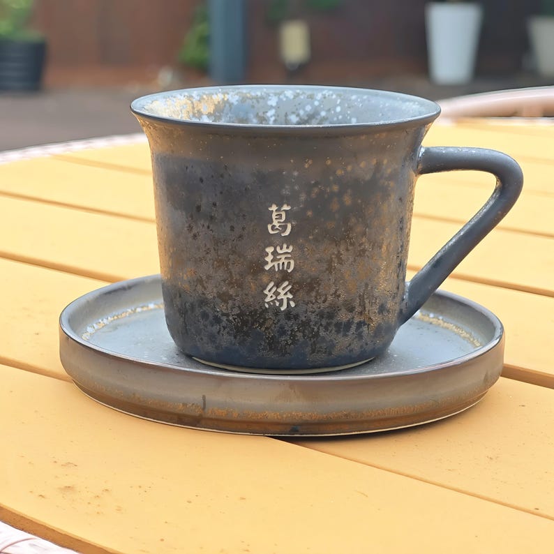 May include: A dark blue ceramic mug with a matching saucer. The mug has a handle and the text "萬瑞絲" in white. The mug and saucer are on a yellow wooden surface, suggesting a rustic or handmade aesthetic.