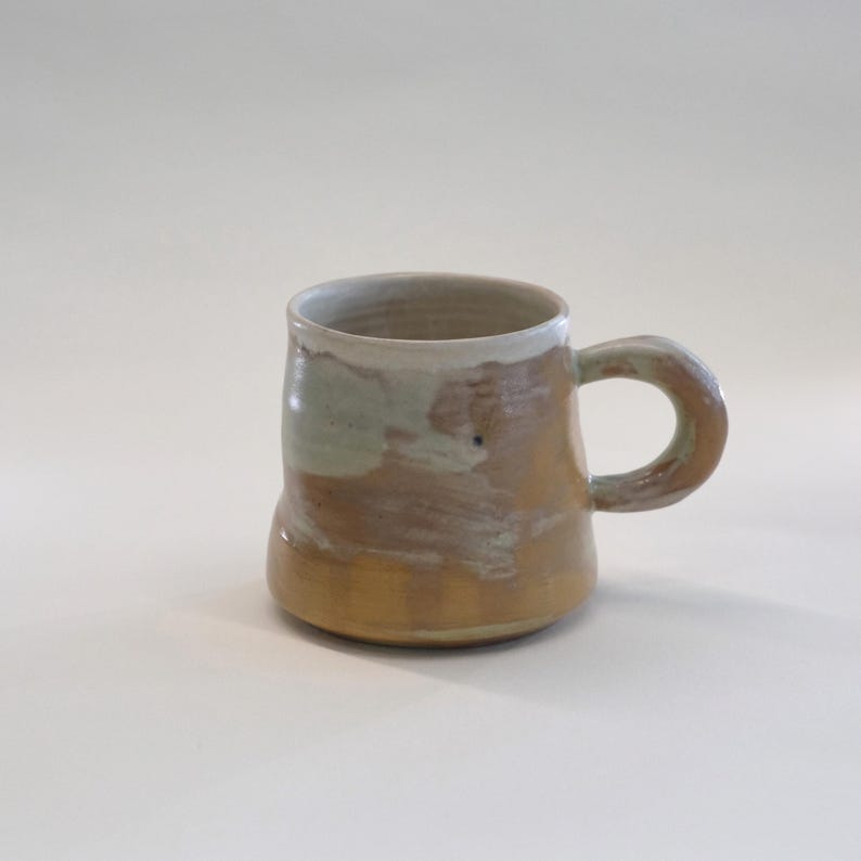 May include: A handmade ceramic mug with a rounded base and a large handle. The mug features a color palette of tan, brown, and off-white, with a touch of green. The mug is suitable for coffee, tea, or other beverages.