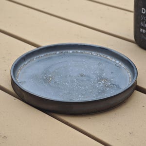 May include: A round, ceramic plate with a dark gray rim and a light gray center. The plate has a speckled pattern and a slightly raised edge. The plate is sitting on a light brown wooden surface.