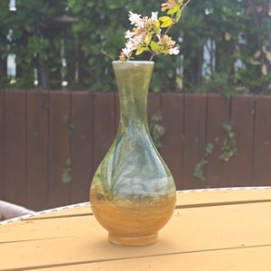 K&ouml;nnte beinhalten: Eine handgefertigte Keramikvase in Gr&uuml;n- und Braunt&ouml;nen. Die Vase hat einen bauchigen Boden und einen schmalen Hals, in dem sich ein Zweig mit wei&szlig;en und gelben Bl&uuml;ten befindet. Die Vase steht auf einem gelben Tisch.