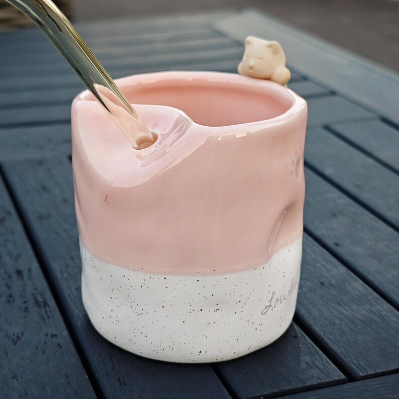 May include: A pink and white ceramic mug with a small cat figurine on the rim. The mug has a speckled white base and a pink upper section. A stream of liquid is pouring into the mug. The word "Louie" is written on the side.