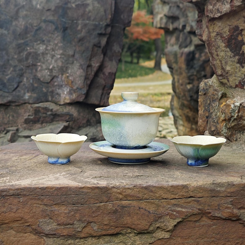 May include: A ceramic tea set with a lidded gaiwan, a saucer, and two small bowls. The set features a cream, blue, and green color scheme. The bowls have a petal-like shape. The set is displayed on a stone surface.