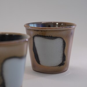 以下が含まれることがあります： 茶色の外観と白と黒の正方形のデザインが施された2つのセラミックカップ。カップは滑らかで光沢のある仕上がりで、シンプルでモダンなデザインです。カップはおそらく手作りで、コーヒーやお茶に適しています。