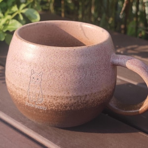 Może przedstawiać: Okrągły ceramiczny kubek z nakrapianym wykończeniem, przechodzącym od jasnego r&oacute;żu u g&oacute;ry do ciemniejszego brązu u podstawy. Kubek posiada mały, wygrawerowany rysunek liniowy kota oraz napis "Home Buddy" nadrukowany na przedniej stronie.
