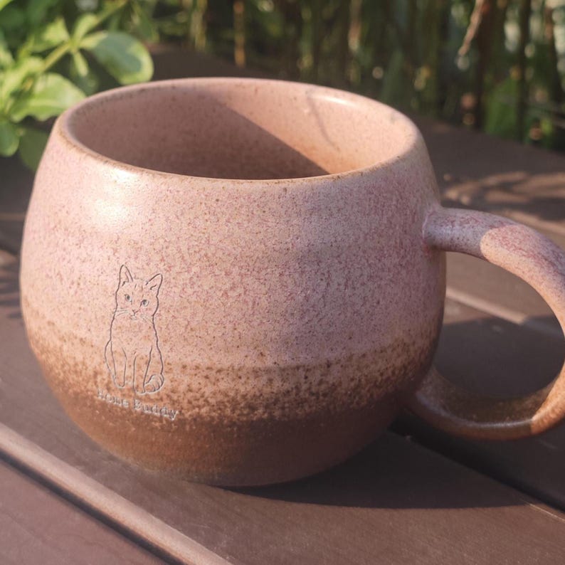 Może przedstawiać: Okrągły ceramiczny kubek z nakrapianym wykończeniem, przechodzącym od jasnego r&oacute;żu u g&oacute;ry do ciemniejszego brązu u podstawy. Kubek posiada mały, wygrawerowany rysunek liniowy kota oraz napis "Home Buddy" nadrukowany na przedniej stronie.