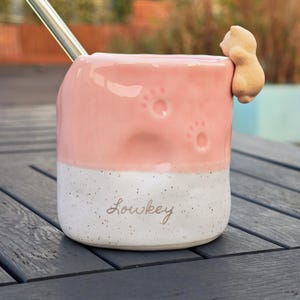 May include: Pink and white ceramic mug with paw prints and a small bear figurine attached. The mug has the word "Lowkey" written on it and a stainless steel straw. A unique and charming design for a beverage cup.
