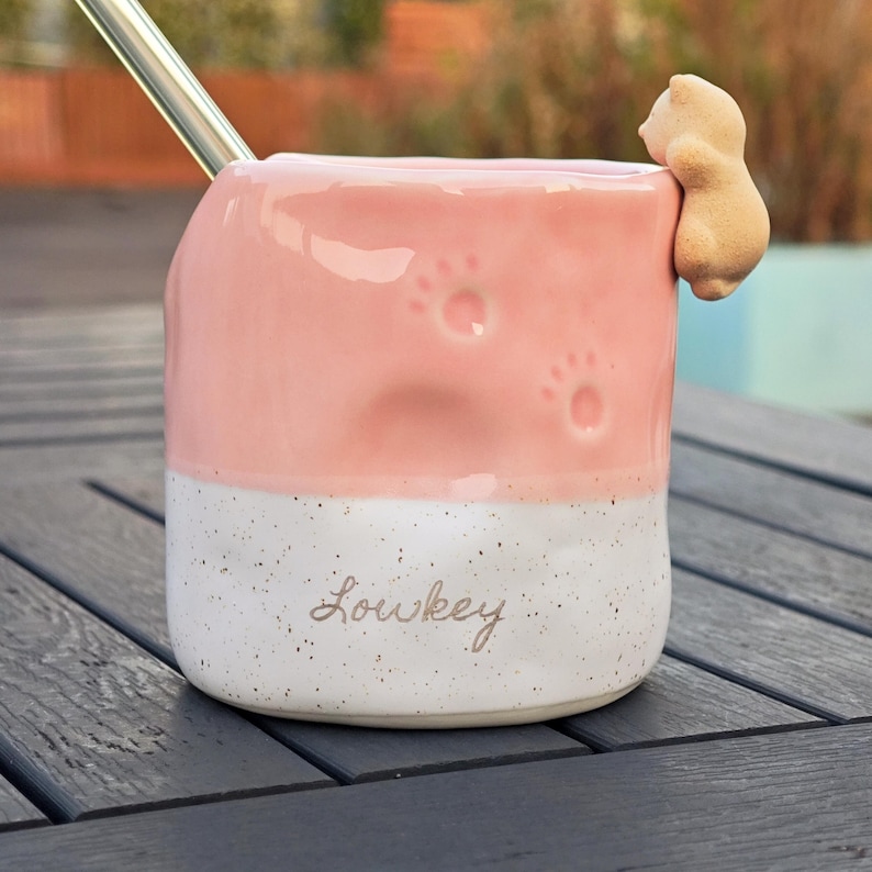 May include: Pink and white ceramic mug with paw prints and a small bear figurine attached. The mug has the word "Lowkey" written on it and a stainless steel straw. A unique and charming design for a beverage cup.