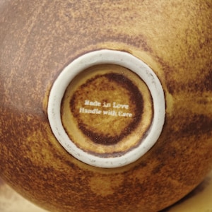 May include: Close-up of the bottom of a brown ceramic bowl with a white rim. The center of the bowl has the text "Made in Love Handle with Care". The bowl has a textured surface.