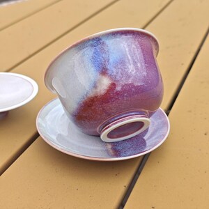 May include: A ceramic teacup and saucer set. The teacup is a vibrant blend of purple, red, and white hues, resting on a matching saucer. The set is placed on a wooden surface, showcasing its elegant design.