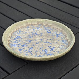 May include: A round ceramic plate with a speckled blue and white pattern on a beige background. The rim is a muted green. The plate is likely handmade and suitable for serving food.